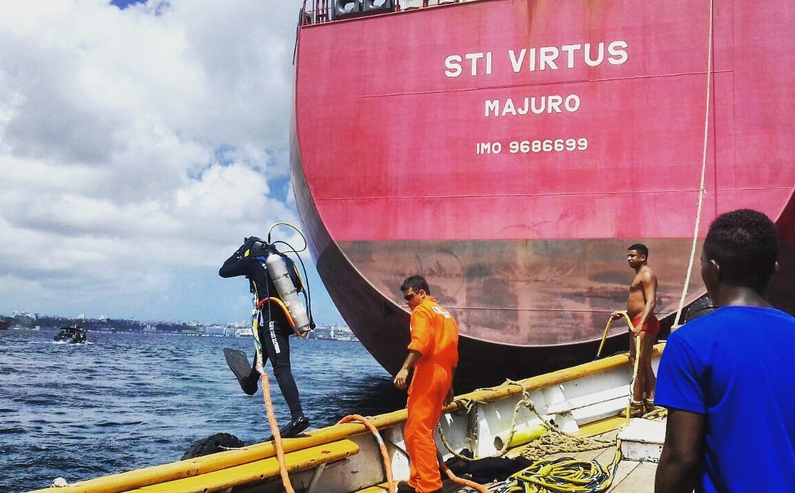 Diver entering water from a boat near a large red ship with several crew members nearby.