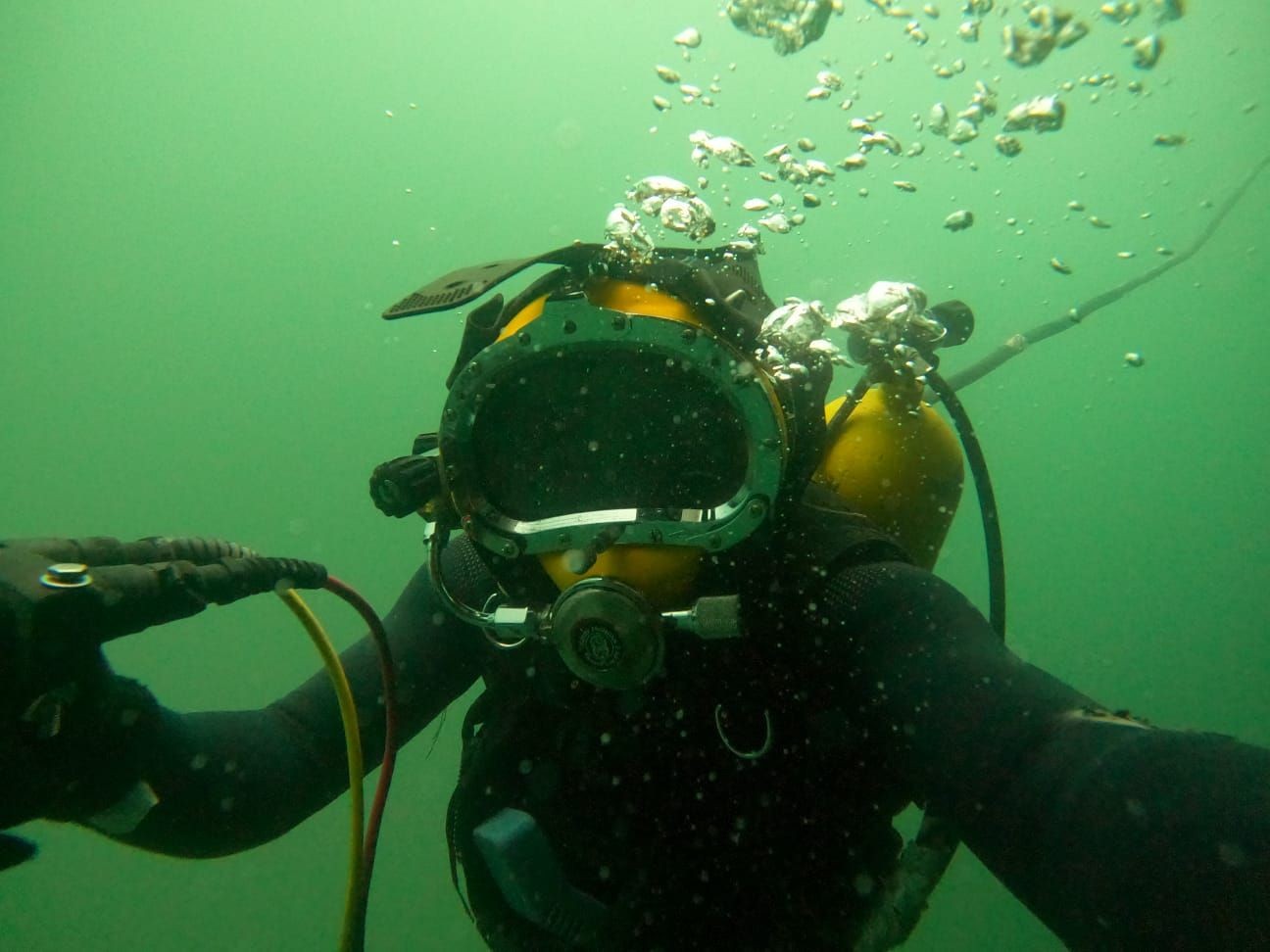 Underwater diver wearing a helmet and breathing apparatus, surrounded by green water and bubbles.