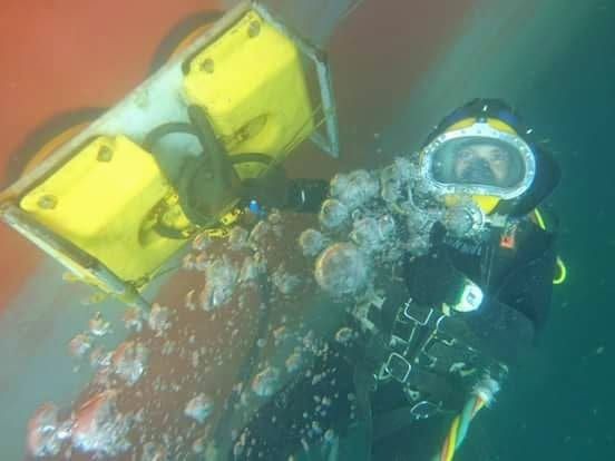 Underwater diver wearing a helmet and suit, operating a yellow mechanical device with bubbles floating around.